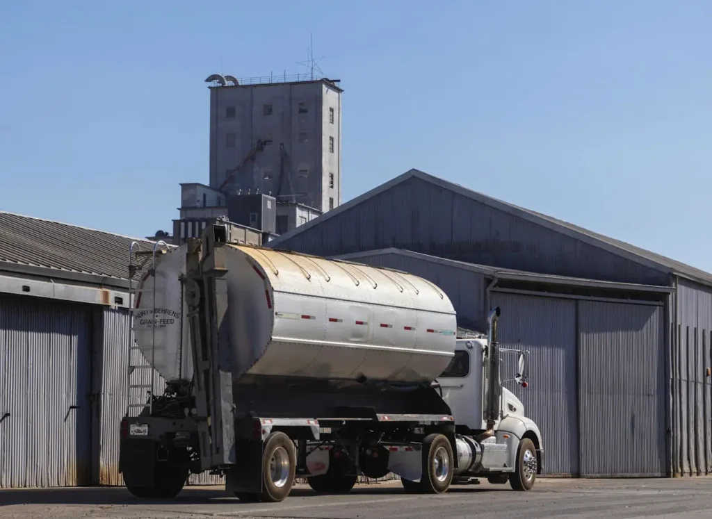 pexels-photo-18468412 Truck transporting a grain tank parked in front of an industrial warehouse with a tall silo in the background under a clear blue sky.